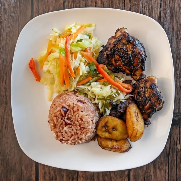 A white plate with jerk chicken, rice and peas, fried plantains, and steamed cabbage.