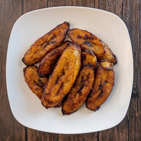 A white plate filled with several golden-brown, grilled plantains on a dark wooden table.