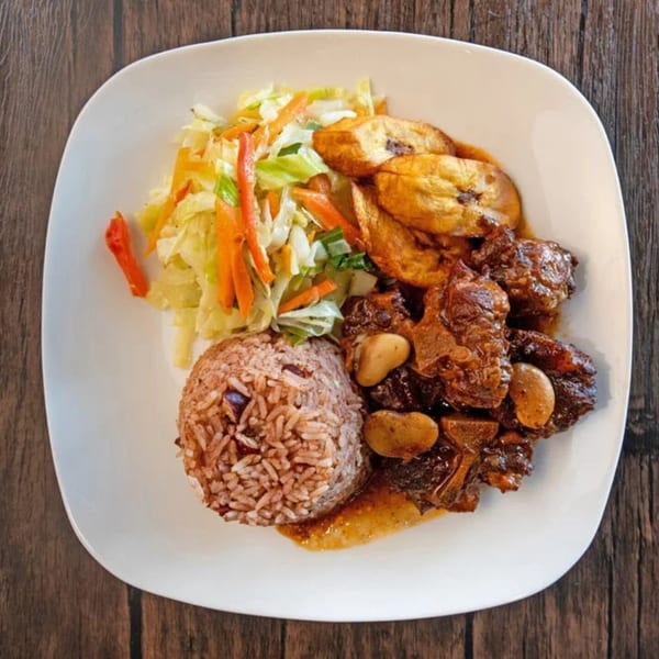 Oxtail stew with rice and peas, fried plantains, and a side salad on a white plate.