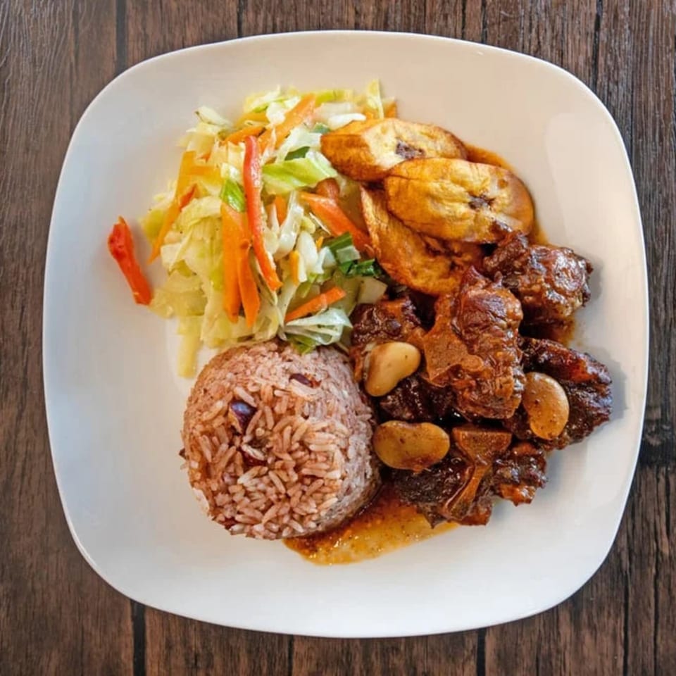 Oxtail stew with rice and peas, fried plantains, and a side salad on a white plate.